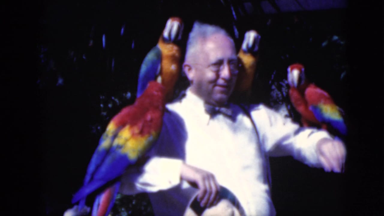 Smiling man in a bow tie with macaws perched on his shoulders and arm.