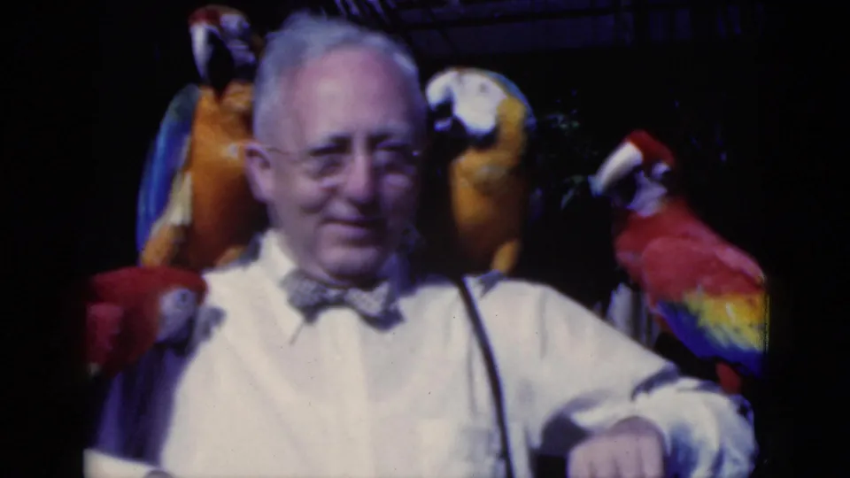 1975 Florida frame of a man in a white shirt and bow tie standing with macaws on both sides in a dim tropical setting.
