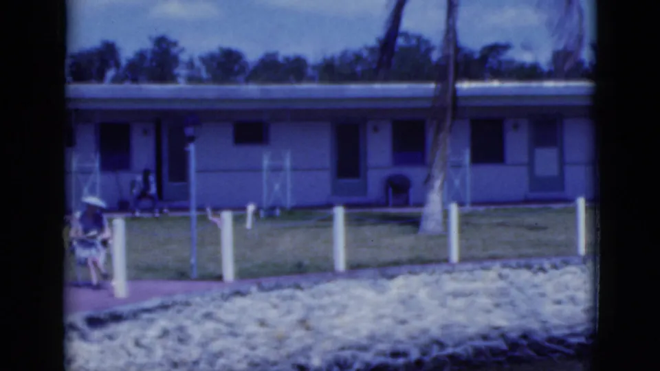 1975 Florida home-movie frame showing a low roadside motel-like exterior with lawn, white fence posts, walkway, and palm trunk.