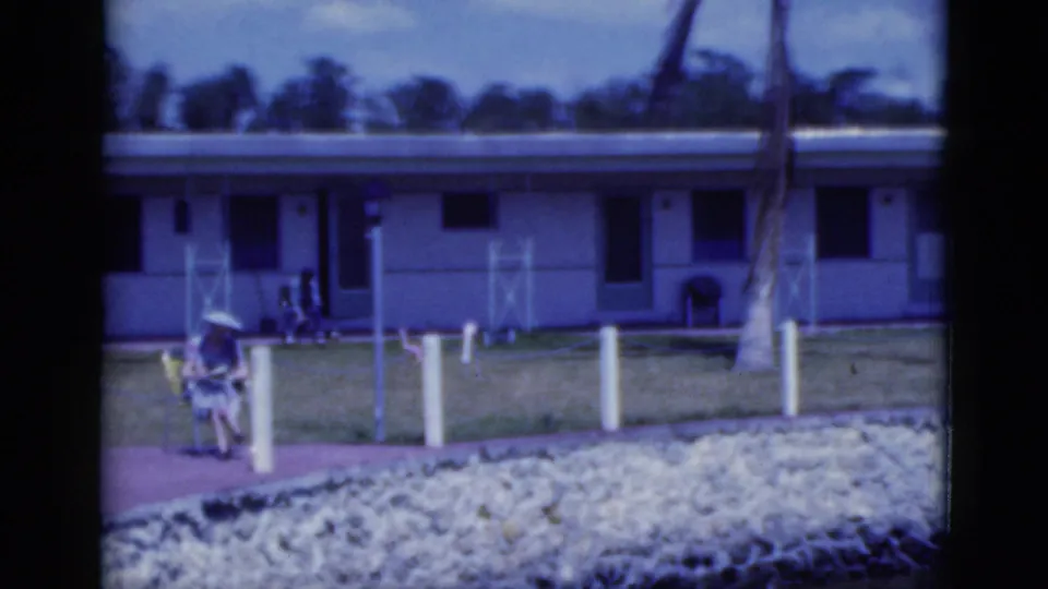 1975 Florida home-movie frame of a woman seated outside a motel or resort building on a sunny day.