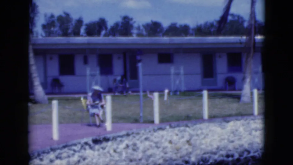 1975 Florida motel or resort scene with a woman seated outdoors near a walkway and palm trees.