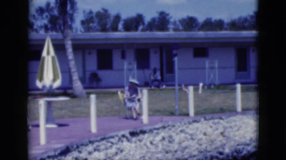 1975 Florida resort or motel frame showing a closed striped umbrella, walkway posts, and a seated woman.