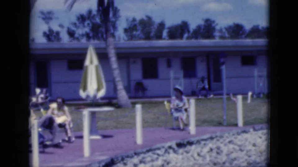 1975 Florida home-movie frame with motel guests outdoors, a closed striped umbrella, palms, and a low resort building.