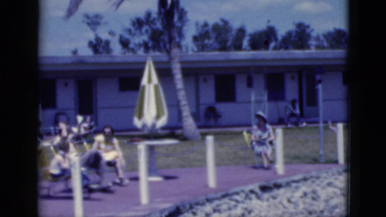 Adults sit in lounge chairs near a striped umbrella outside a low motel-style building.