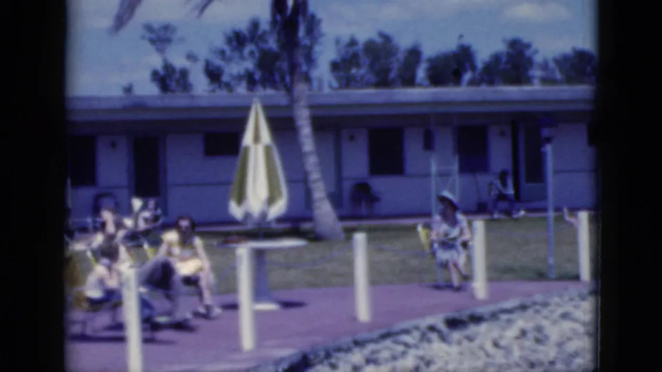 1975 Florida 8mm frame showing adults relaxing by a pool area with a striped umbrella, palm tree, and low motel-style building.