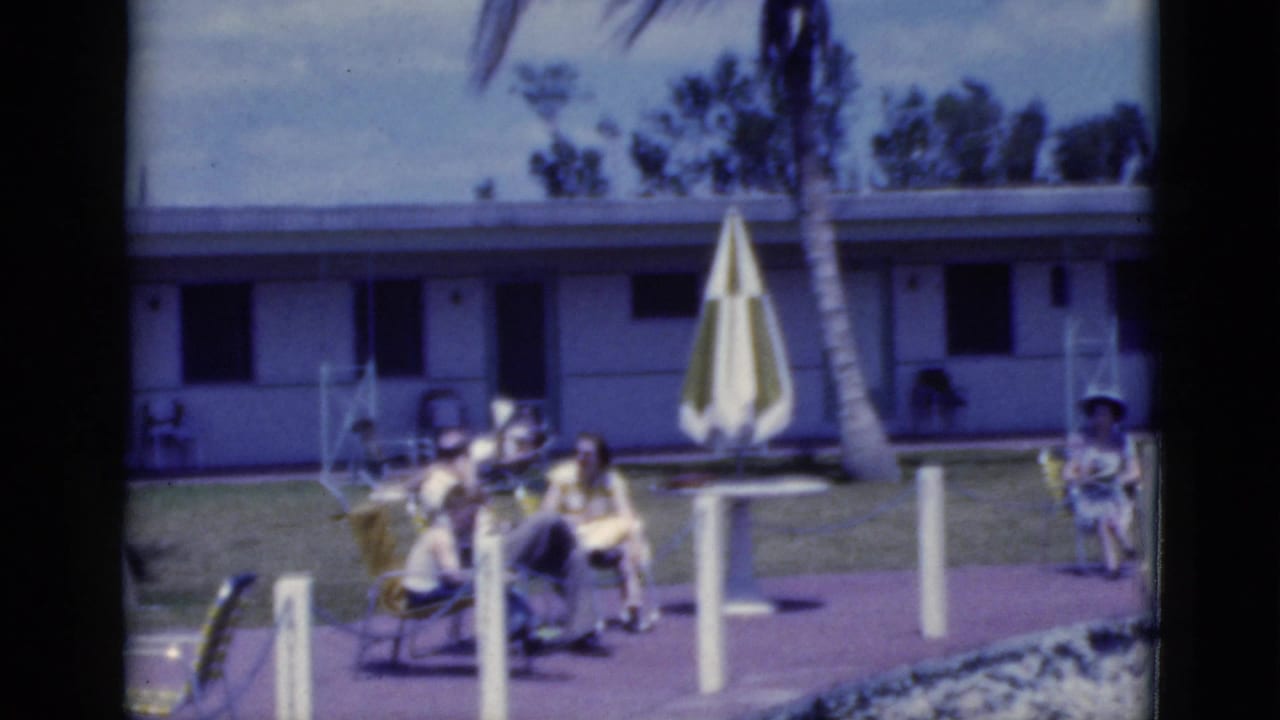 Adults recline in poolside chairs with a striped umbrella and palm tree in front of a low building.