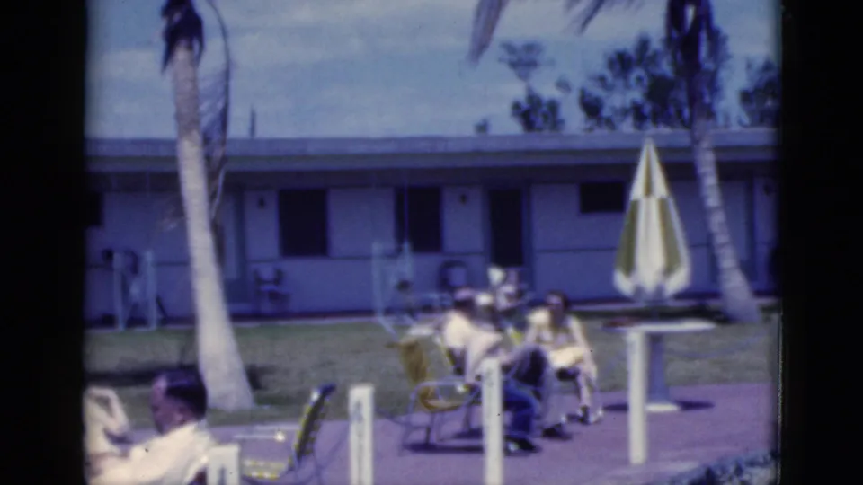 1975 Florida 8mm frame with palm trees, a striped umbrella, and adults seated near a motel-style pool area.