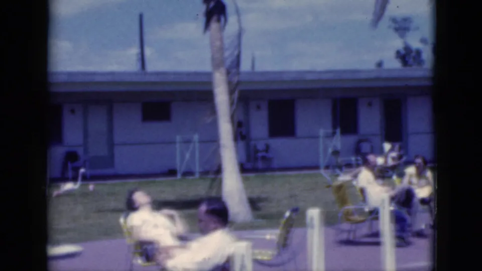 1975 Florida home-movie frame showing adults sunning near a pool patio, with palm trees and a low motel-style building in back.