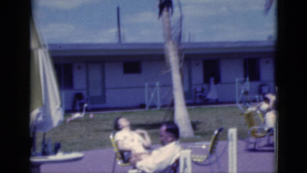 1975 Florida 8mm frame showing adults seated outdoors near lawn chairs, a palm tree, grass, patio, and a low motel-style building.
