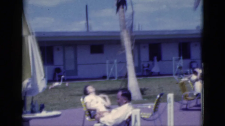 1975 Florida 8mm frame showing adults seated outdoors near lawn chairs, a palm tree, grass, patio, and a low motel-style building.