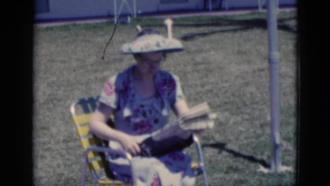 1975 Florida home-movie frame of a woman in a floral dress and broad sun visor seated on grass in a striped lawn chair with a newspaper.