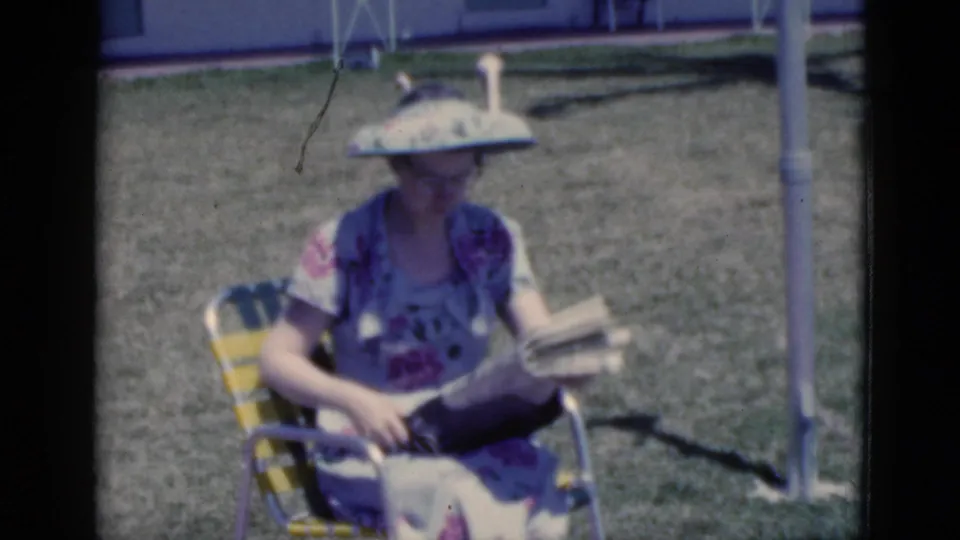 1975 Florida home-movie frame of a woman in a floral dress and broad sun visor seated on grass in a striped lawn chair with a newspaper.