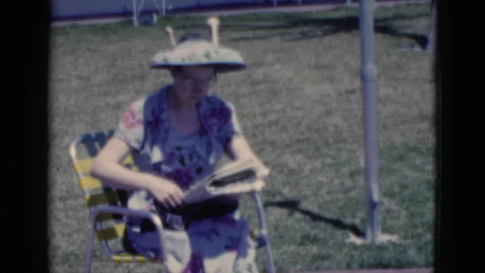 1975 Florida 8mm frame showing a woman in a floral dress and large visor seated in a yellow-green lawn chair with an open newspaper.