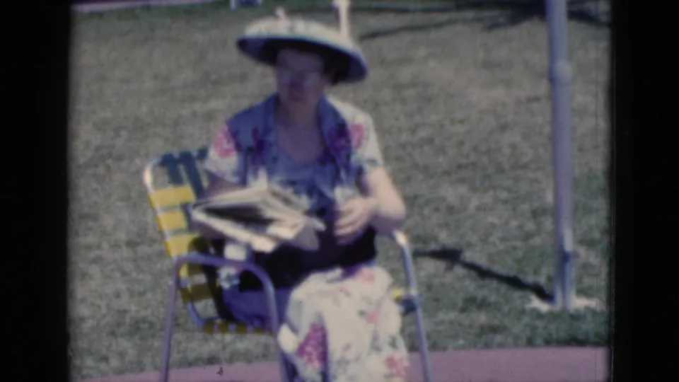 1975 Florida home-movie frame of a woman seated in a striped lawn chair, wearing a broad sun visor and floral dress while reading a newspaper.