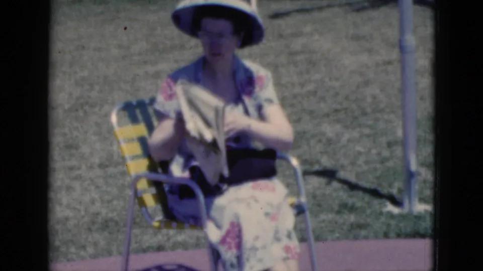 In 1975 Florida home-movie footage, a woman in a floral dress and sun hat reads a newspaper in a yellow and green striped lawn chair.