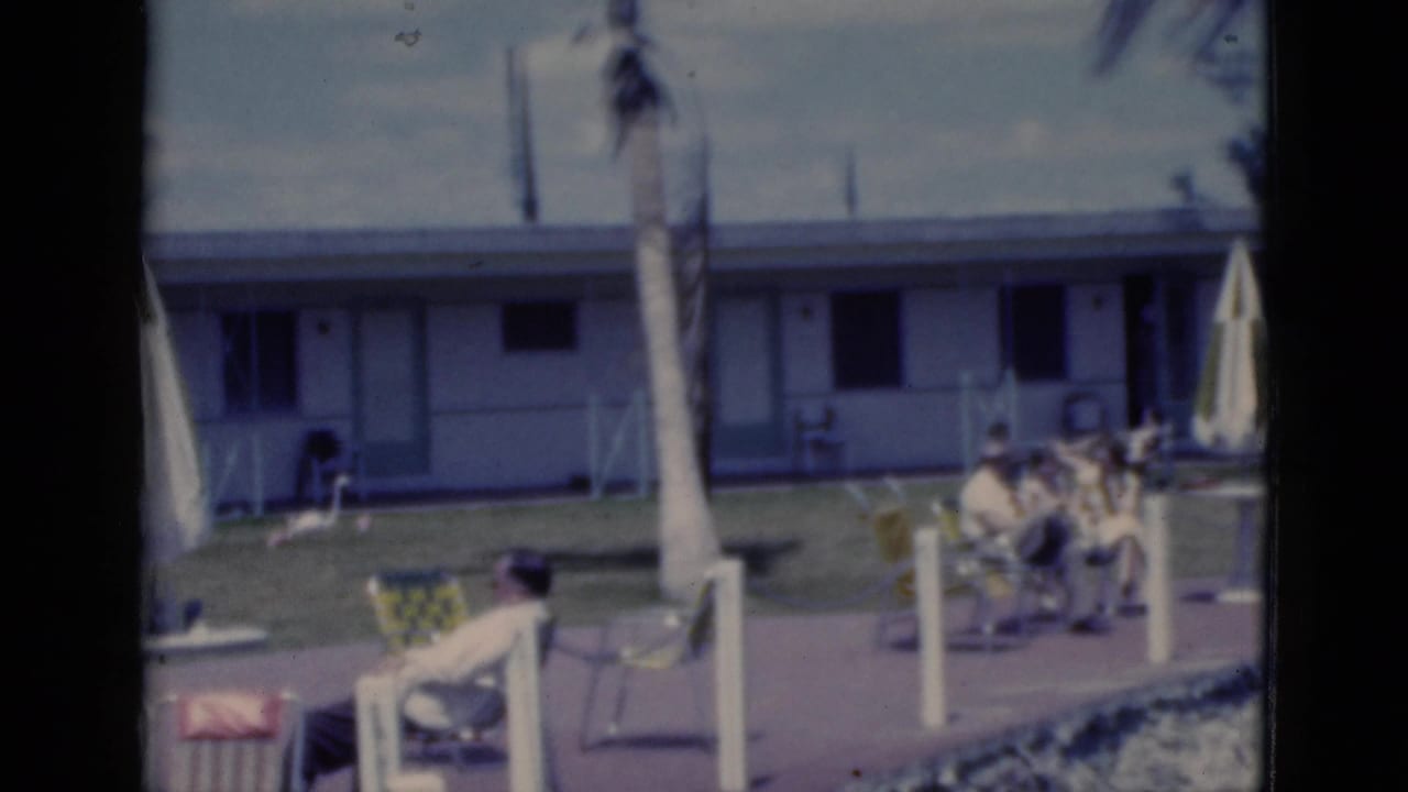 Adults relaxing in patio chairs outside a low motel building with palm trees and umbrellas.
