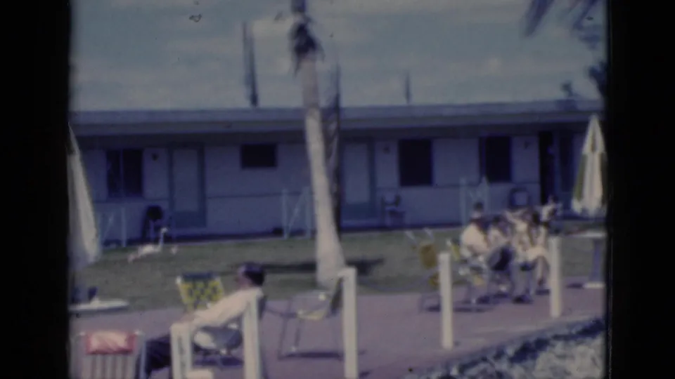 1975 Florida home-movie frame showing adults seated on a sunny motel patio with lounge chairs, closed umbrellas, palms, and rooms behind them.
