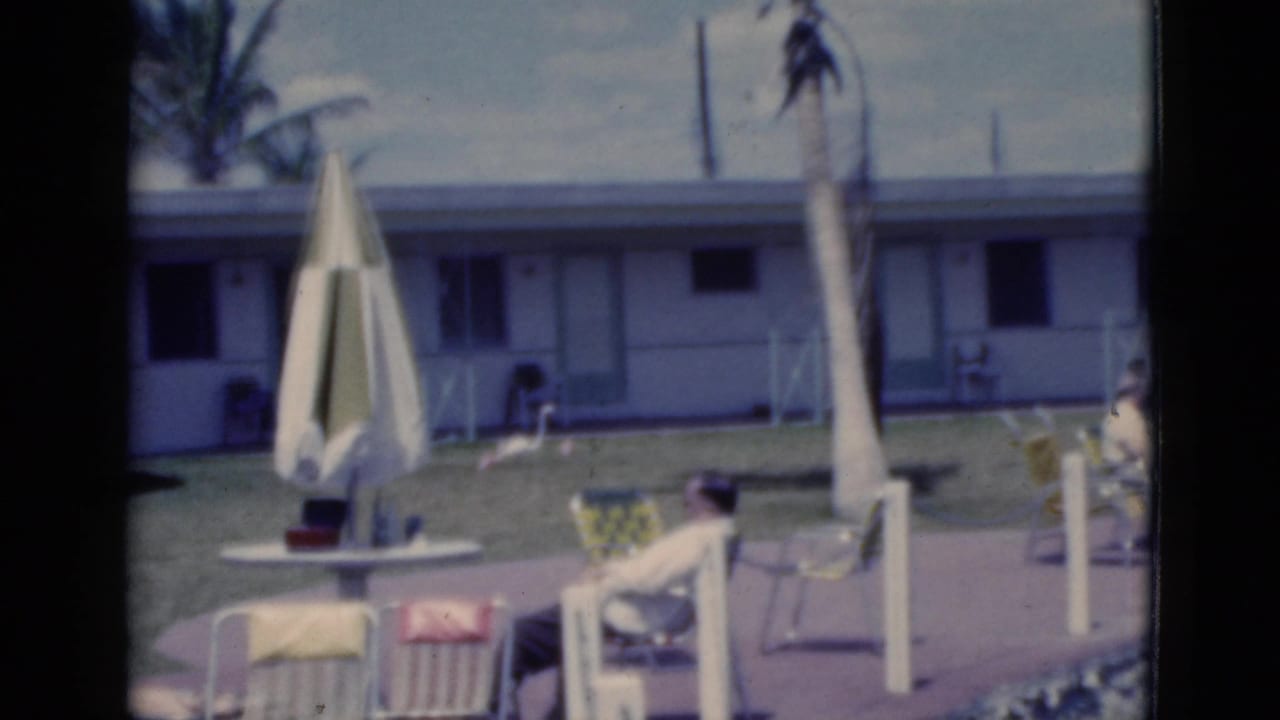 Man seated in a lounge chair near a closed patio umbrella outside motel rooms.