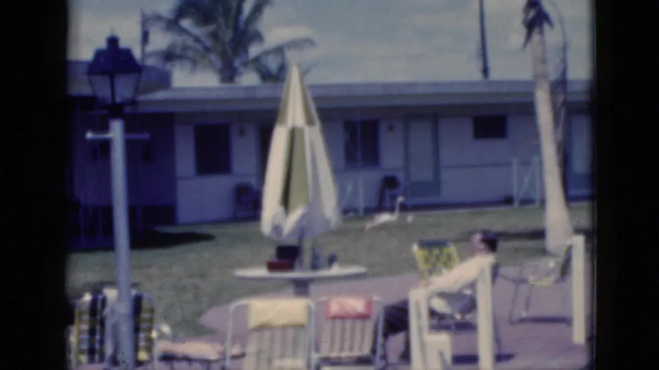 1975 Florida home-movie frame showing a patio area with a closed umbrella, seated guest, lamppost, palm trees, lawn, and low motel building.