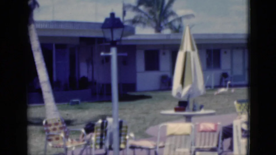 1975 Florida home-movie frame of a sunny motel patio with a seated adult, closed umbrella, lamppost, palm trees, lawn, and rooms behind.