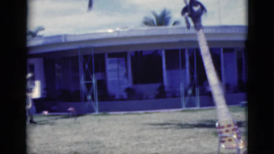 1975 Florida 8mm frame showing a woman in a light outfit near a palm tree and single-story house, with an alligator partly visible below.
