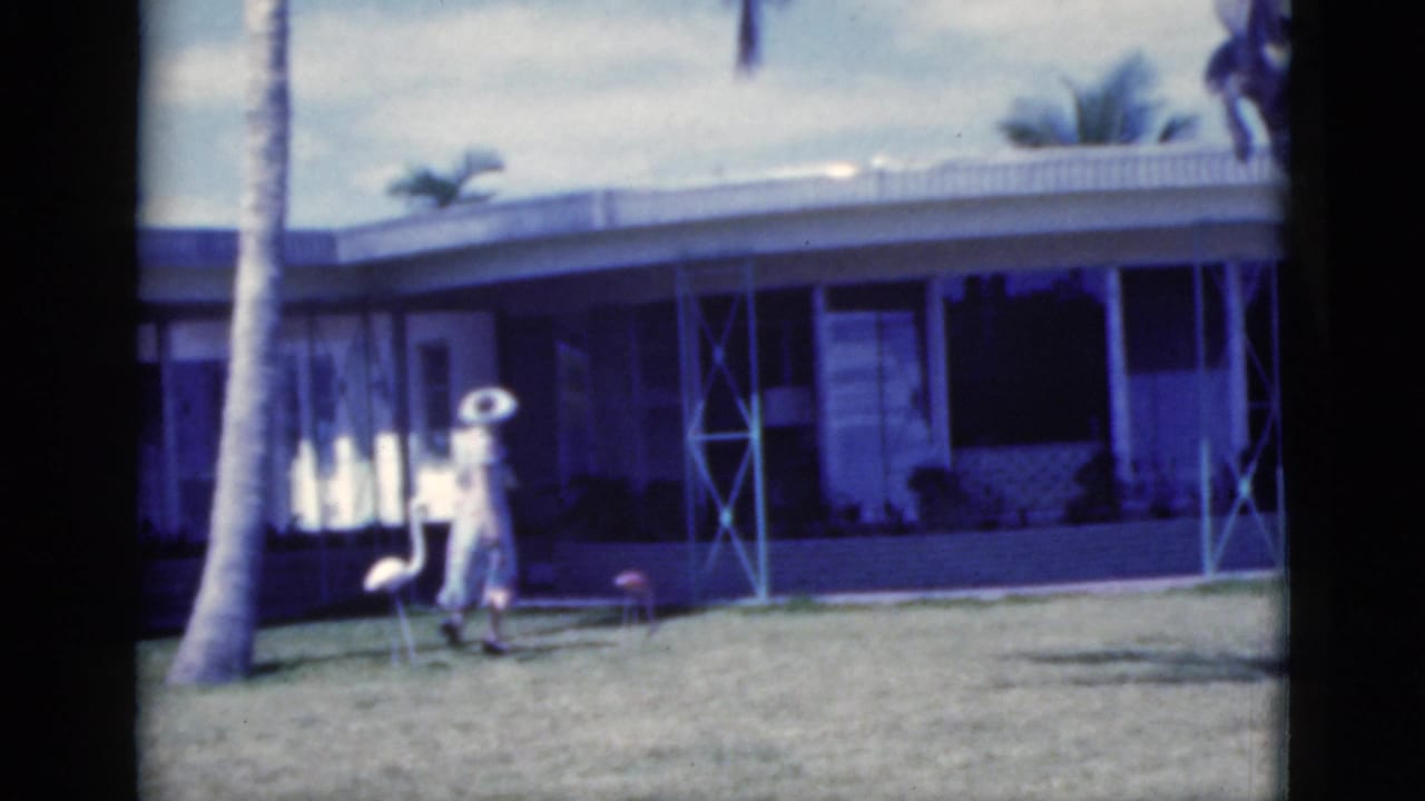 1975 Florida home-movie frame of a woman in a light dress and wide-brimmed hat walking across a lawn before a low house.