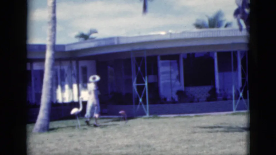 1975 Florida home-movie frame of a woman in a light dress and wide-brimmed hat walking across a lawn before a low house.