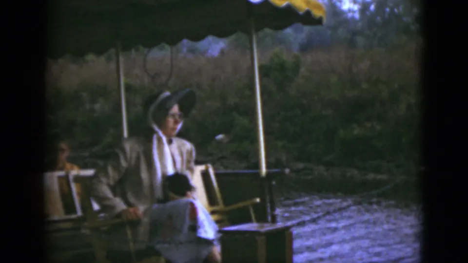 A woman in a hat, glasses, light suit, and floral dress sits on a covered Florida riverboat in 1975.