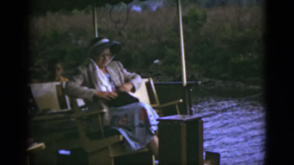 The woman sits quietly on the covered riverboat, holding a book or bag on her lap as water passes beside the deck.