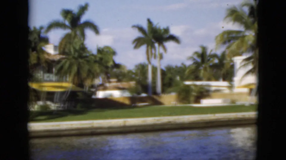 1975 Florida home-movie frame showing canal-front homes, lawns, palms, and water viewed from a boat.