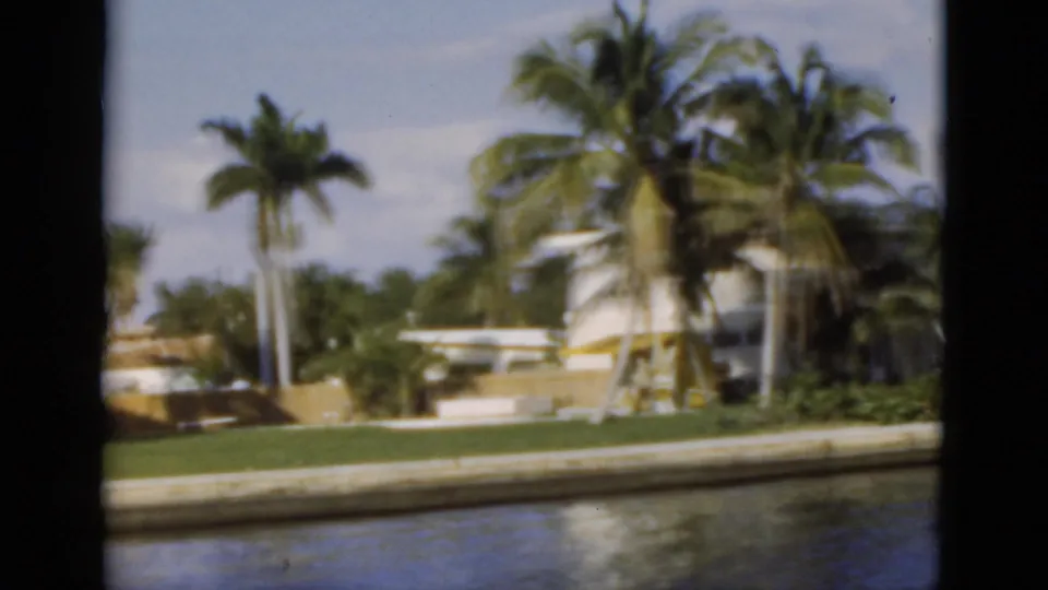 1975 Florida shoreline view with tall palms, a white residential building, lawn, seawall, and water in the foreground.