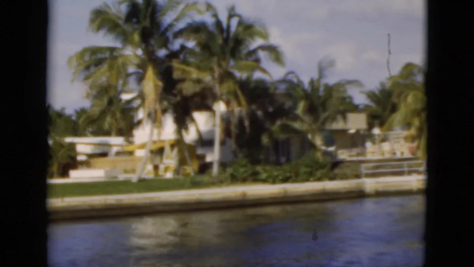 1975 Florida home-movie frame showing a palm-covered canal edge with residential structures and a low seawall.