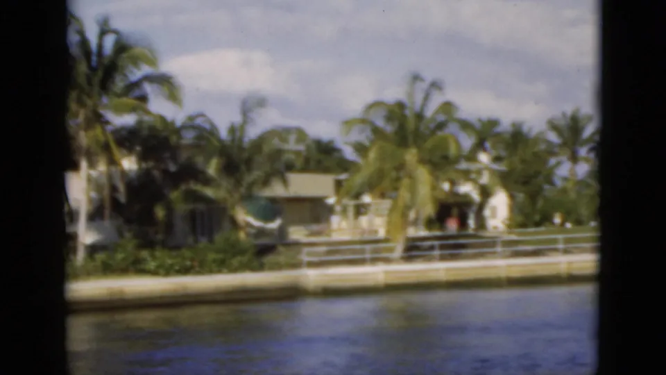 1975 Florida waterfront view with a residence, palms, metal railing, seawall, reflective canal water, and a possible distant person.