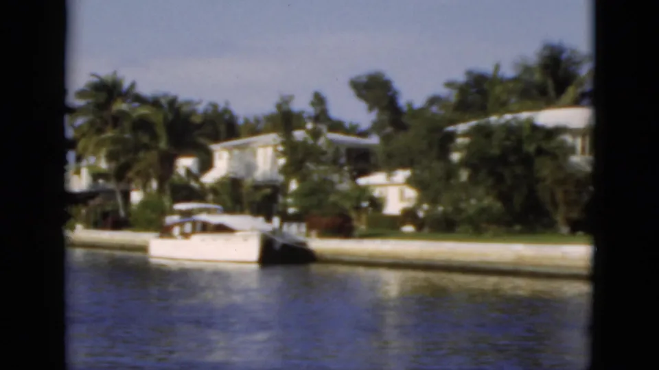 1975 Florida 8mm frame showing canal water, white waterfront homes, palms, seawall, and a small boat near the bank.
