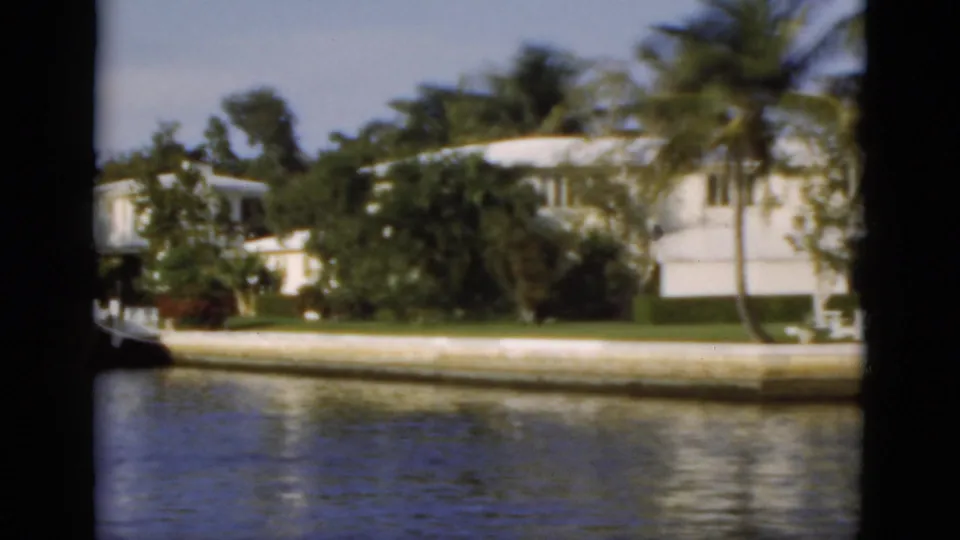 1975 Florida canal scene with white houses, palms, seawall, lawn, and rippling water viewed from a passing boat.