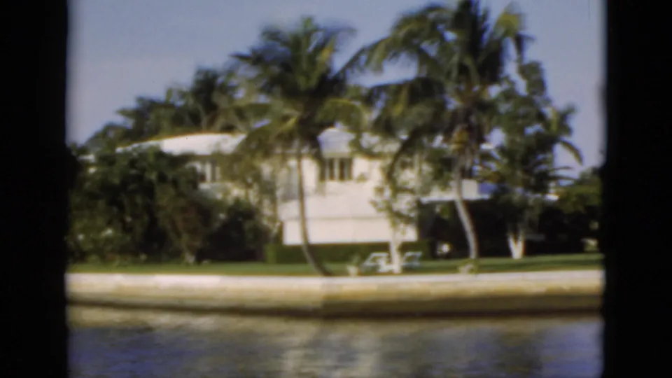 1975 Florida 8mm frame focused on a white waterfront house, palms, lawn chairs, seawall, and canal water.