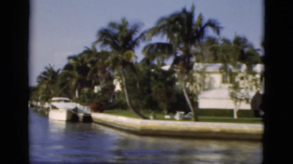 1975 Florida canal frame showing a bend in the waterway, palm-lined homes, seawall, and a small white boat.