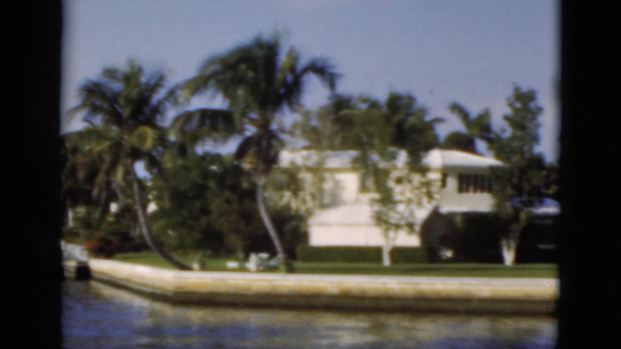 Canal water in front of a white waterfront house, lawn, palms, and dark film-gate borders.