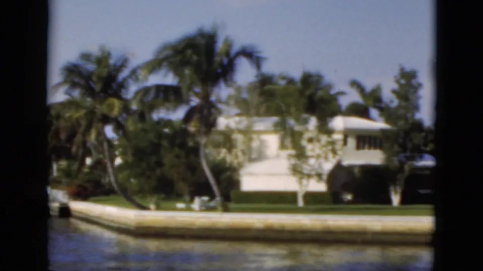 1975 Florida 8mm frame showing a white waterfront home behind a seawall, with canal water, palms, lawn, and dark gate edges.