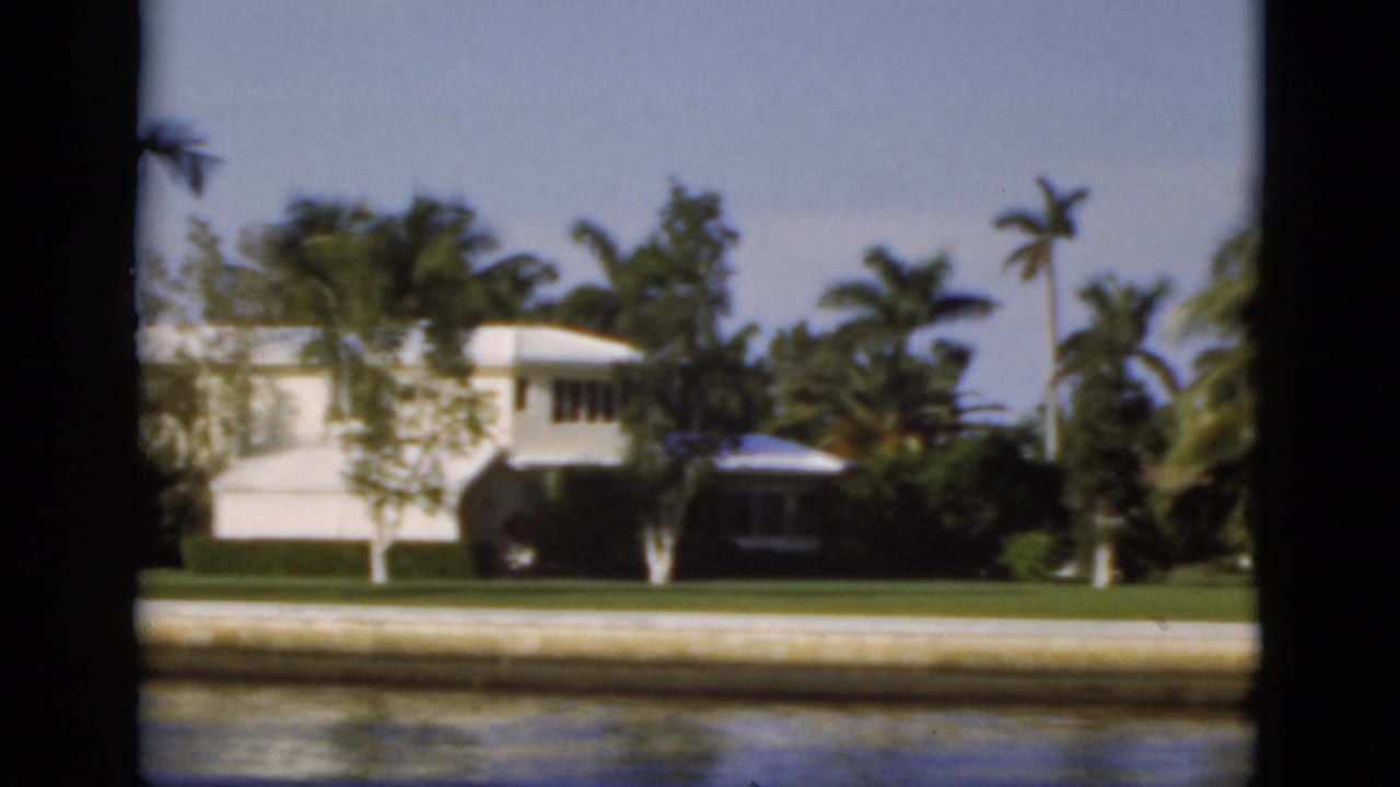 White waterfront house and small docked boat seen across a canal with palms and lawn.
