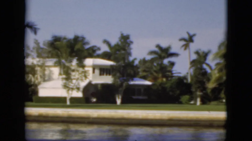 1975 Florida canal view with a white waterfront house, small docked boat, seawall, palms, lawn, and dark frame borders.