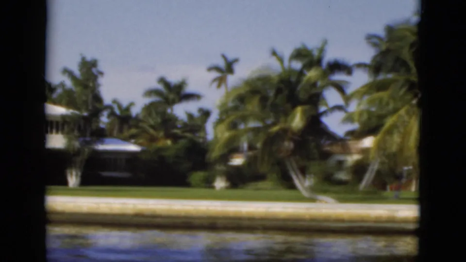 1975 Florida 8mm frame showing a palm-lined canal edge, partial white house, lawn, seawall, and small docked boat.
