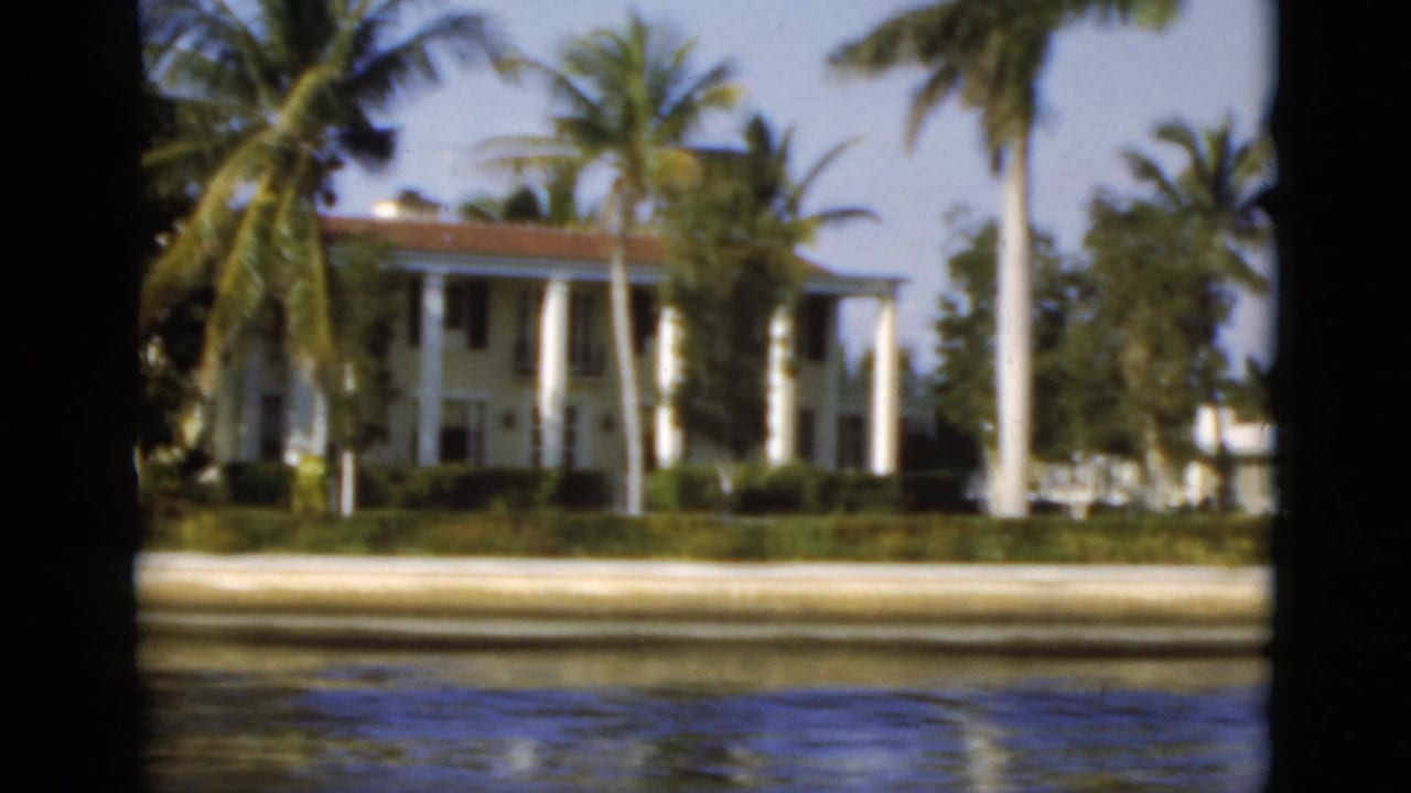 A large columned waterfront house in Florida appears behind palms and a seawall in the 1975 home-movie footage.