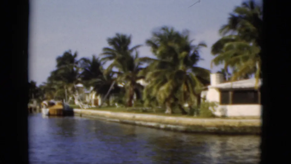 1975 Florida canal scene with palms, calm water, seawalls, docks, and residential buildings along the bank.