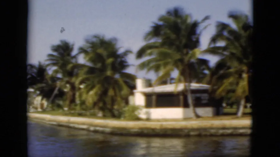 1975 Florida 8mm frame showing a white canal-front home, palms, seawall, lawn, and water from a passing boat.