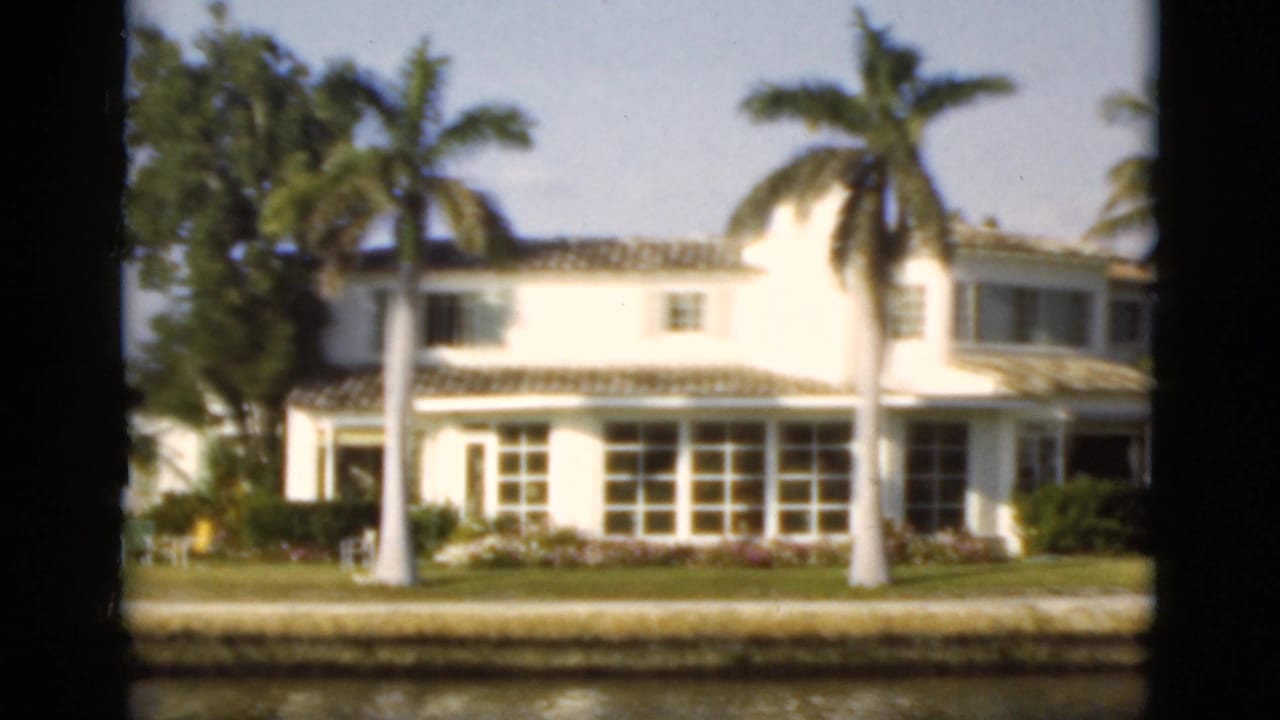 1975 Florida home-movie frame of a white canal-side house with tiled roof, palms, screened windows, lawn, and seawall.