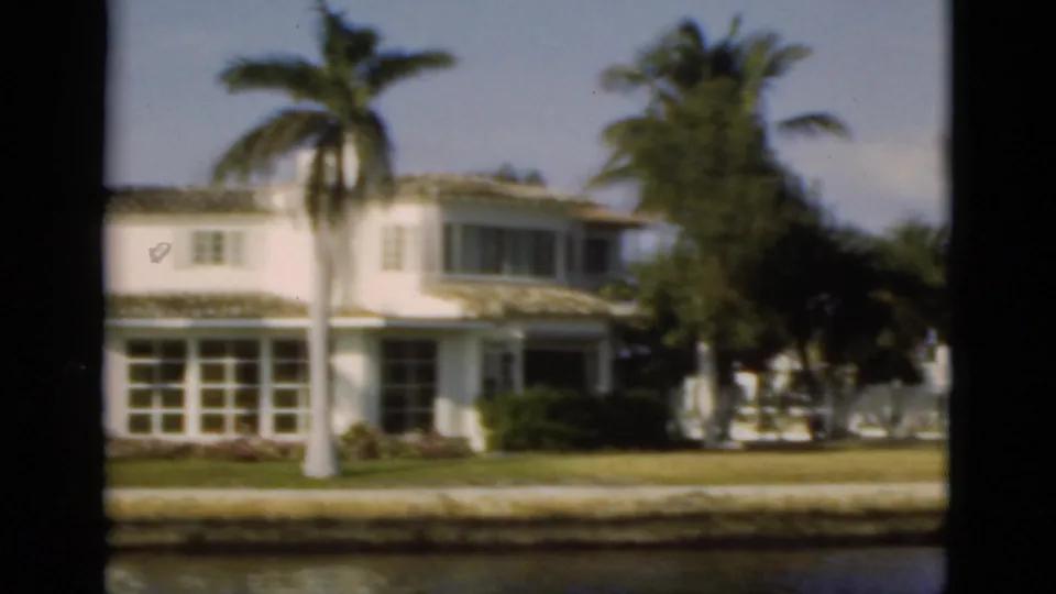 1975 Florida 8mm frame showing a white two-story waterfront home, palm trunk, screened windows, landscaping, and canal edge.