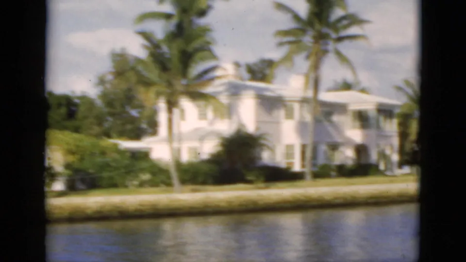 1975 Florida home-movie frame of another white canal-side residence with palms, shrubs, lawn, seawall, and reflective water.