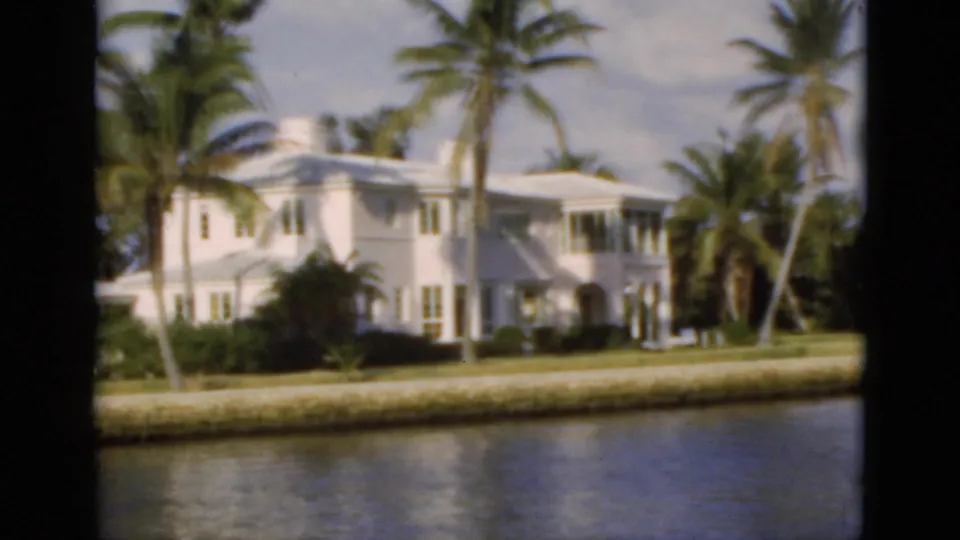 1975 Florida 8mm frame showing a large white waterfront house, palms, sea wall, and calm water viewed from across the channel.
