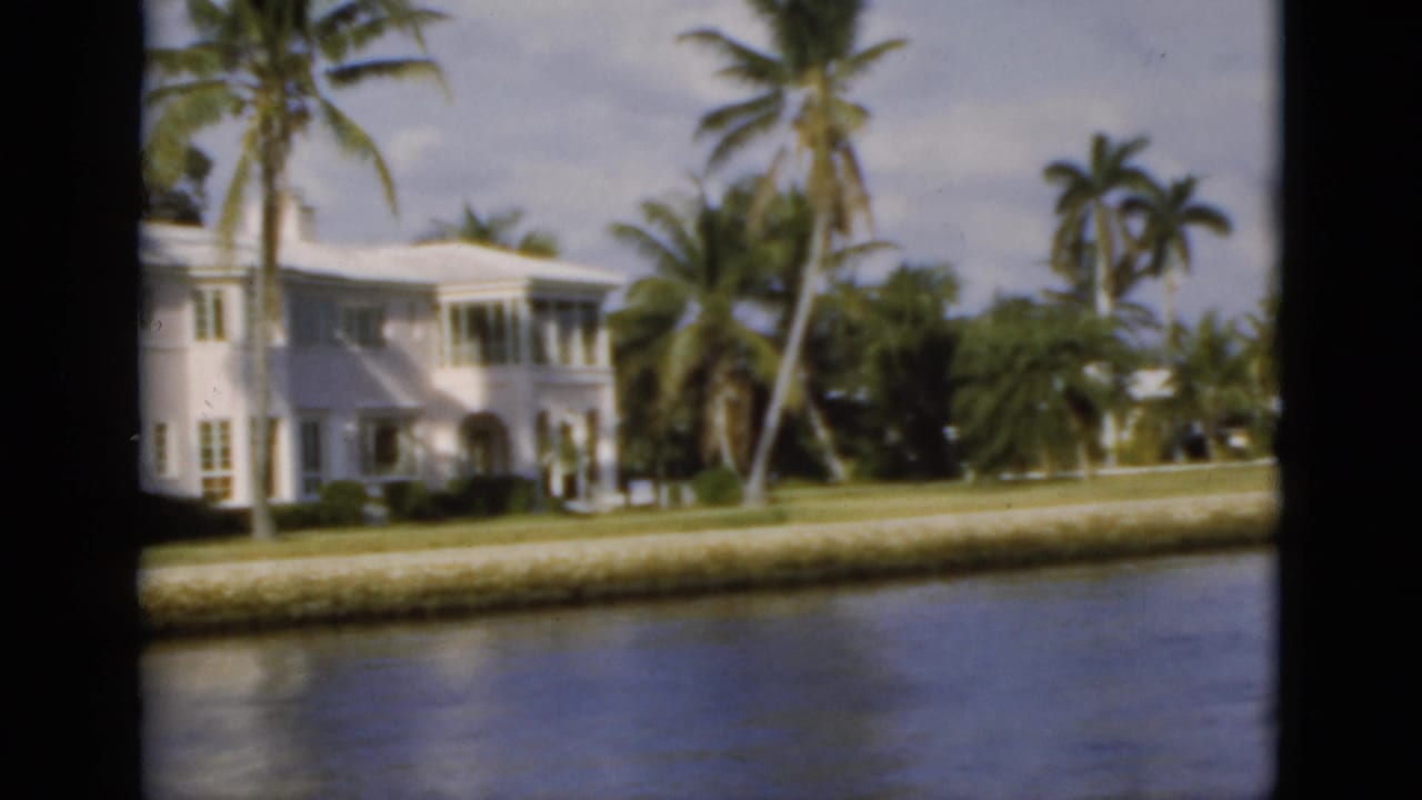 White shoreline house with tall palms, lawn, sea wall, and water in the foreground.